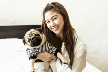 Sticker - Beautiful Asian young woman playing with her dog and smile with dog pug breed looking in funny face in bedroom,Friendship Concept