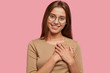 canvas print picture - Generous thankful friendly looking girl keeps hands on heart, has good attitude, dressed in casual clothes, isolated over pink background. Joyful kind lady feels thankfulness. Horizontal view