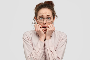 Anxious freckled girl looks nervously at camera, stares in distrust, holds breath with anticipation of horror, bites finger nails, dressed in fashionable shirt, isolated on white studio wall