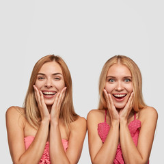 Vertical portrait of joyful two girls with broad smiles, being amazed as see something incredible and nice, stand next to each other, isolated over white background. Positive emotions concept