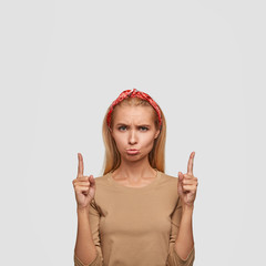 Dissatisfied blonde woman with offensive look, gloomy expression, points with both index fingers upwards, dressed in beige casual sweater, poses against white background. Its very unpleasant!