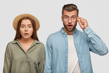 Wall Mural - Wow! Surprised lovers stare at camera with full disbelief, hear shocking news, stand next to each other. Beautiful terrified woman in straw hat and unshaven guy being in stupor, isolated on white