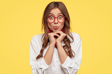 Poster - European teenager has amazed look at camera, keeps hands under chin, wears spectacles, white shirt, bandana poses over yellow background, pouts lips, has surprised facial expression. Youth concept