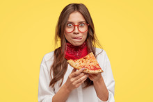 Pretty Woman With Appealing Appearance, Licks Lips, Holds Slice Of Tasty Pizza, Being Hungry, Wants To Eat, Enjoys Taste, Has Good Appetite, Isolated Over Yellow Studio Wall. People And Snack Concept