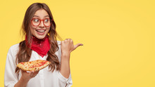 Fastfood And Meal Concept. Cheerful Young Girl Smiles Pleasantly, Eats Tasty Slice Of Pizza, Being In Good Mood, Shows Place She Bought It, Advertises Pizzeria, Isolated Over Yellow Background.