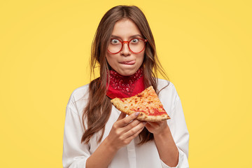 Wall Mural - Pretty woman with appealing appearance, licks lips, holds slice of tasty pizza, being hungry, wants to eat, enjoys taste, has good appetite, isolated over yellow studio wall. People and snack concept