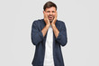 © WHstudio Leushin N - Dejected astonished displeased man frowns face, keeps hands on cheeks, regrets something, has negative facial expression, wears white t shirt and blue shirt, poses against white studio wall.