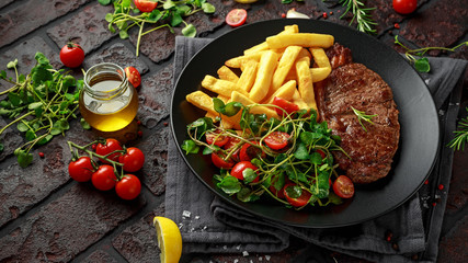 Wall Mural - Grilled sirloin steak with potato fries and vegetables, tomato salad in a black plate. rustic table