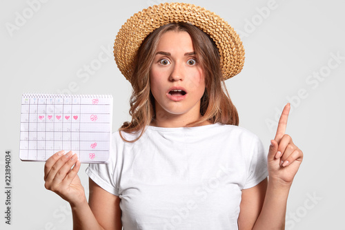 Fotografie, Obraz Shocked European woman with terrified expression, raises index finger, wears straw hat and casual t shirt, holds periods calendar, isolated over white background
