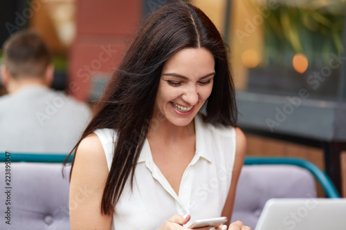 Fotografie, Tablou Outdoor shot of pleased brunette woman with appealing look, uses modern gadget, recieves pleasant notification from friend, texts feedback, has rest in cafeteria, wears white blouse