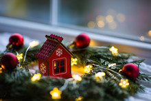 Red Wooden Toy House Surrounded With Fir-tree Wreath Decorated With Warm Garland Lights And Little Christmas Balls Near Window.