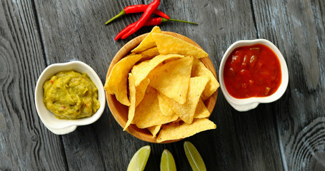 Flat lay of bowl with corn nacho chips served on wooden table with different sauces and chili pepper