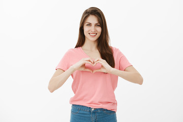 Show some love to closest one. Portrait of happy cute and gently young female in pink t-shirt and jeans, showing heart gesture over chest and smiling broadly, showing loving and passionate feelings