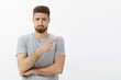 © Cookie Studio - Sad and cute charming bearded male model in grey t-shirt frowning making gloomy face with frowned eyebrows, sulking pointing right expressing regret and envy standing unhappy over white background