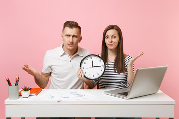 Two young fun shocked business woman man colleagues sit work at white desk with clock, laptop isolated on pastel pink background. Achievement career concept. Copy space advertising, youth co working.