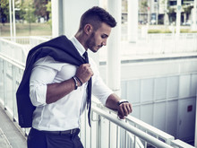 Portrait Of Stylish Young Man Wearing Business Suit, Standing In Modern City Setting, Looking At Wrist Watch To Check The Time