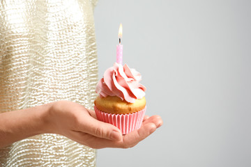 Woman holding delicious birthday cupcake with burning candle on light background, closeup