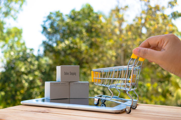 Hand holding trolley and stack of cardboard boxes on tablet with nature background. Consumer can buy products anywhere anytime from seller using web browser. Online shopping and e-commerce concept.