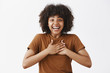 Surprised and greatful attractive tender african american woman in trendy brown t-shirt holding hands on heart and smiling broadly from amazement and joy happy receive award over gray wall