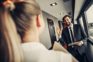 Wall Mural - I am glad to see you. Smiling man gesticulating hand while holding clipboard in hand. He greeting with female colleague