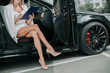 © Yakobchuk Olena - Happy businesswoman making notes in document while locating in contemporary vehicle with opened door. She wearing attractive shoes and throwing leg on another one