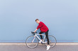 © bodnarphoto - Happy young man in a red sweatshirt sitting on a white bike against a blue wall background. Stylish smiling cyclist on a blue background. Copyspace