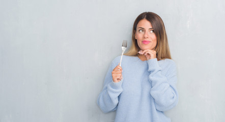 Canvas Print - Young adult woman over grey grunge wall holding silver fork serious face thinking about question, very confused idea