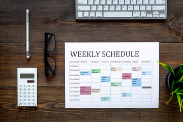 Weekly schedule. Planning the week. Multicolored planner on manager, marketologist's dark wooden office desk with computer keyboard and calculator top view