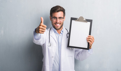 Wall Mural - Handsome young doctor man over grey grunge wall holding clipboard happy with big smile doing ok sign, thumb up with fingers, excellent sign