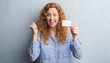 © Krakenimages.com - Young redhead woman over grey grunge wall holding blank visit card screaming proud and celebrating victory and success very excited, cheering emotion