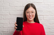 © Krakenimages.com - Young Chinese woman over brick wall showing smartphone with a happy face standing and smiling with a confident smile showing teeth