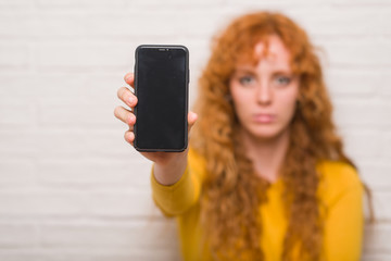 Wall Mural - Young redhead woman sitting over brick wall showing smartphone with a confident expression on smart face thinking serious