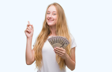 Sticker - Blonde teenager woman holding dollars surprised with an idea or question pointing finger with happy face, number one