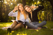 © Sunrise Team - Happy young beautiful ladies students sitting in the park outdoors on grass holding copybook doing homework showing okay gesture.