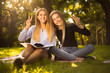 © Sunrise Team - Young beautiful ladies students sitting in the park outdoors on grass holding copybook doing homework showing peace gesture.
