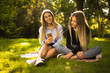 © Sunrise Team - Happy cheerful smiling girls sisters students sitting in the park outdoors on grass using mobile phone.
