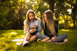 © Sunrise Team - Shocked surprised girls sisters students sitting in the park outdoors on grass using mobile phone listening music with earphones.