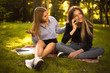 © Sunrise Team - Displeased confused girl looking at her friend sister talking by mobile phone students sitting in the park outdoors on grass.