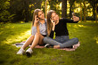© Sunrise Team - Happy girls sisters students sitting in the park outdoors on grass have a rest take a selfie by mobile phone showing okay gesture.