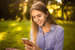 © Sunrise Team - Happy laughing girl student sitting in the park outdoors on grass have a rest using mobile phone.