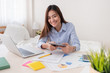 © weedezign - Asian freelancer looking at camera and holding tablet while working with laptop on table in bedroom at home.Work at home concept.work from home concept.