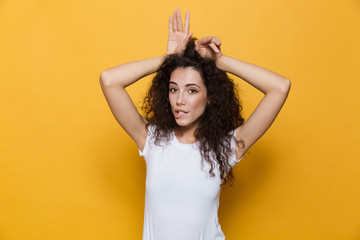 Wall Mural - Image of flirting european woman 20s with curly hair showing rabbit ears at her head, isolated over yellow background