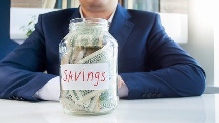 Concept of successful money saving. Young businessman with glass jar for savings full of money