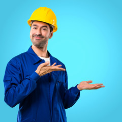 Young workman with helmet extending hands to the side and smiling for presenting and inviting to come on blue background