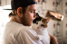 Close-up Of Beard Man In Icelandic Sweater Who Is Holding And Kissing His Cute Purring Devon Rex Cat. Muzzle Of A Cat And A Man's Face. Love Cats And Humans. Relationship, Weasel.
