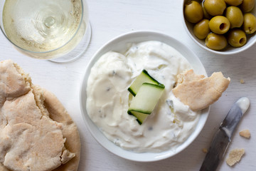 Tzatziki in white ceramic bowl with a cucumber slice and a piece of pita bread next to pita bread, olives and a glass of white wine from above.