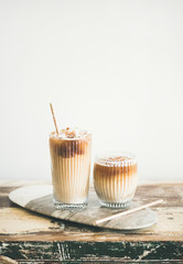 Iced coffee in tall glasses with milk and straws on board over wooden table, white wall at background, copy space. Summer refreshing beverage ice coffee drink concept