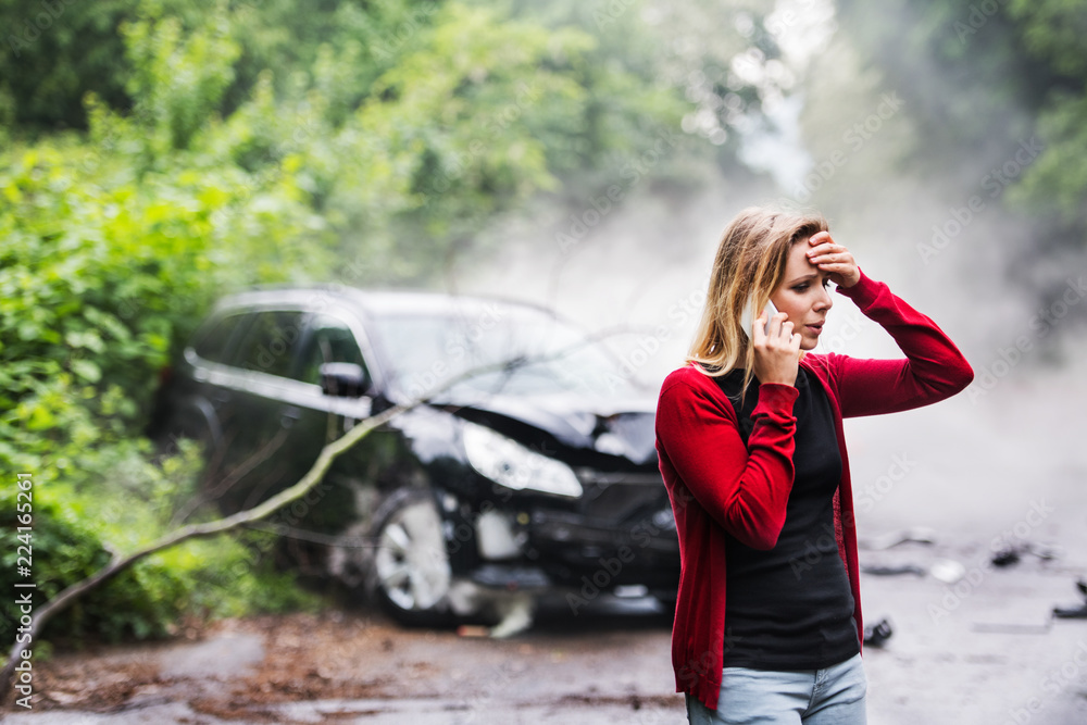 A young woman with smartphone by the damaged car after a car accident, making a phone call. - obrazy, fototapety, plakaty 