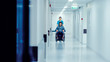 © Gorodenkoff - Female Nurse Pushing Patient in the Wheelchair Through the Hospital Corridor, They are Going to the Procedures. Bright Modern Hospital with Friendly Staff.