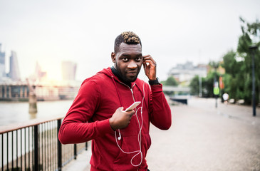 Wall Mural - Young sporty black man runner on the bridge outside in a city, using smartphone.